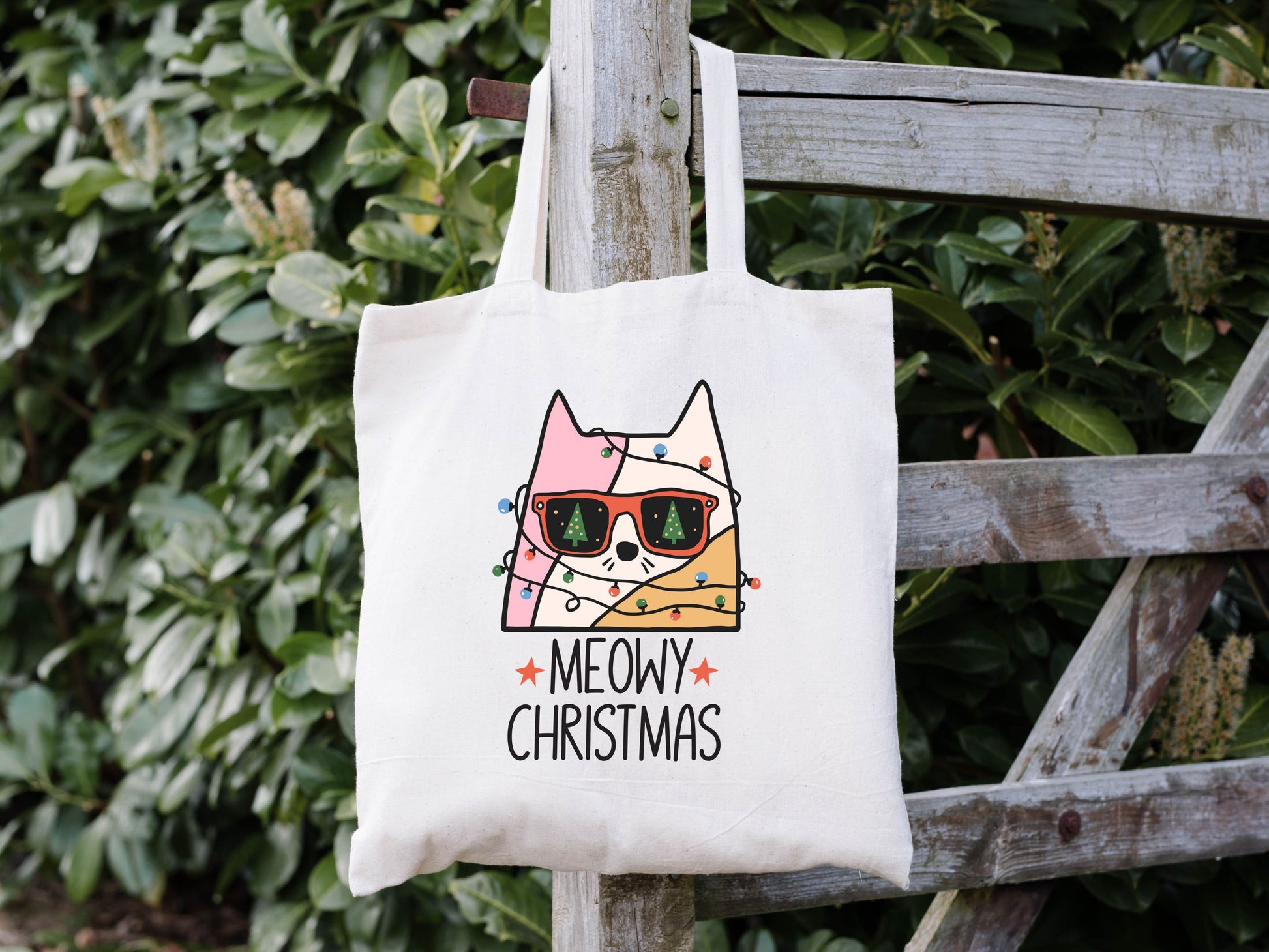 15x15 white tote bag that says "meowy christmas" with an image of a cat wrapped in christmas lights and wearing sunglasses. you can see christmas trees in the reflection of the glasses.