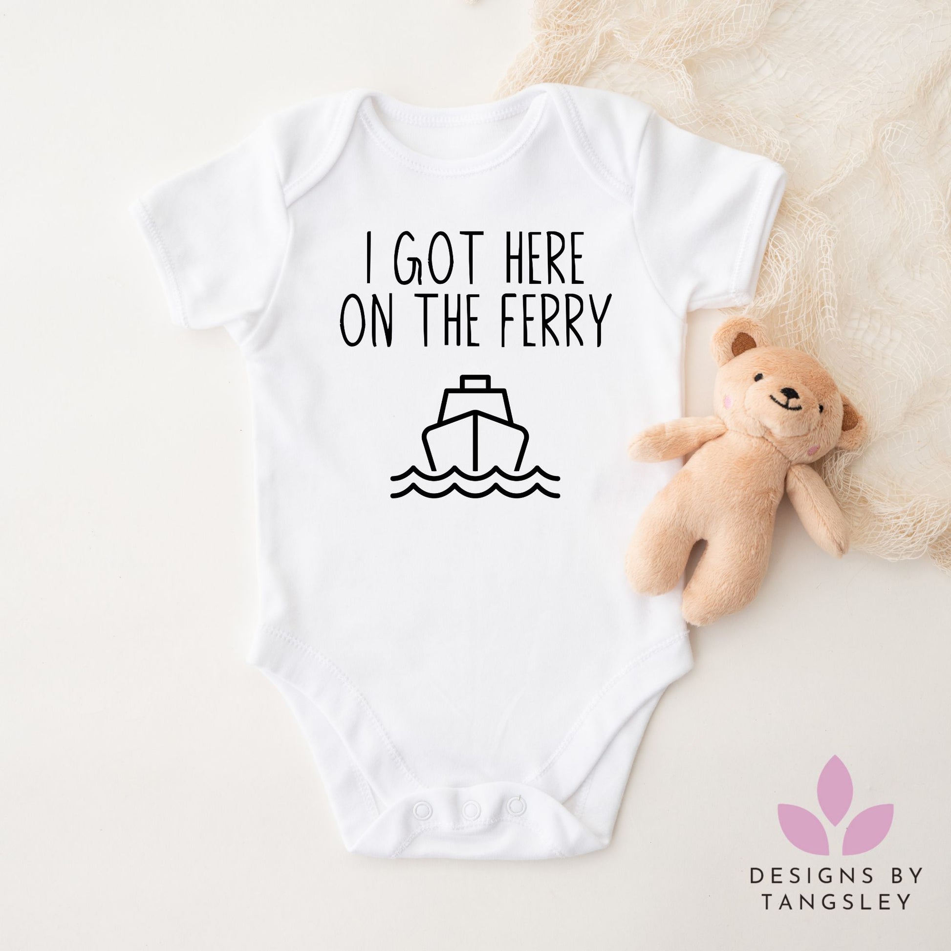 White baby bodysuit with black text "I Got Here On The Ferry" and a simple ferry graphic. Displayed flat with a plush teddy. Handmade by Designs by Tangsley.