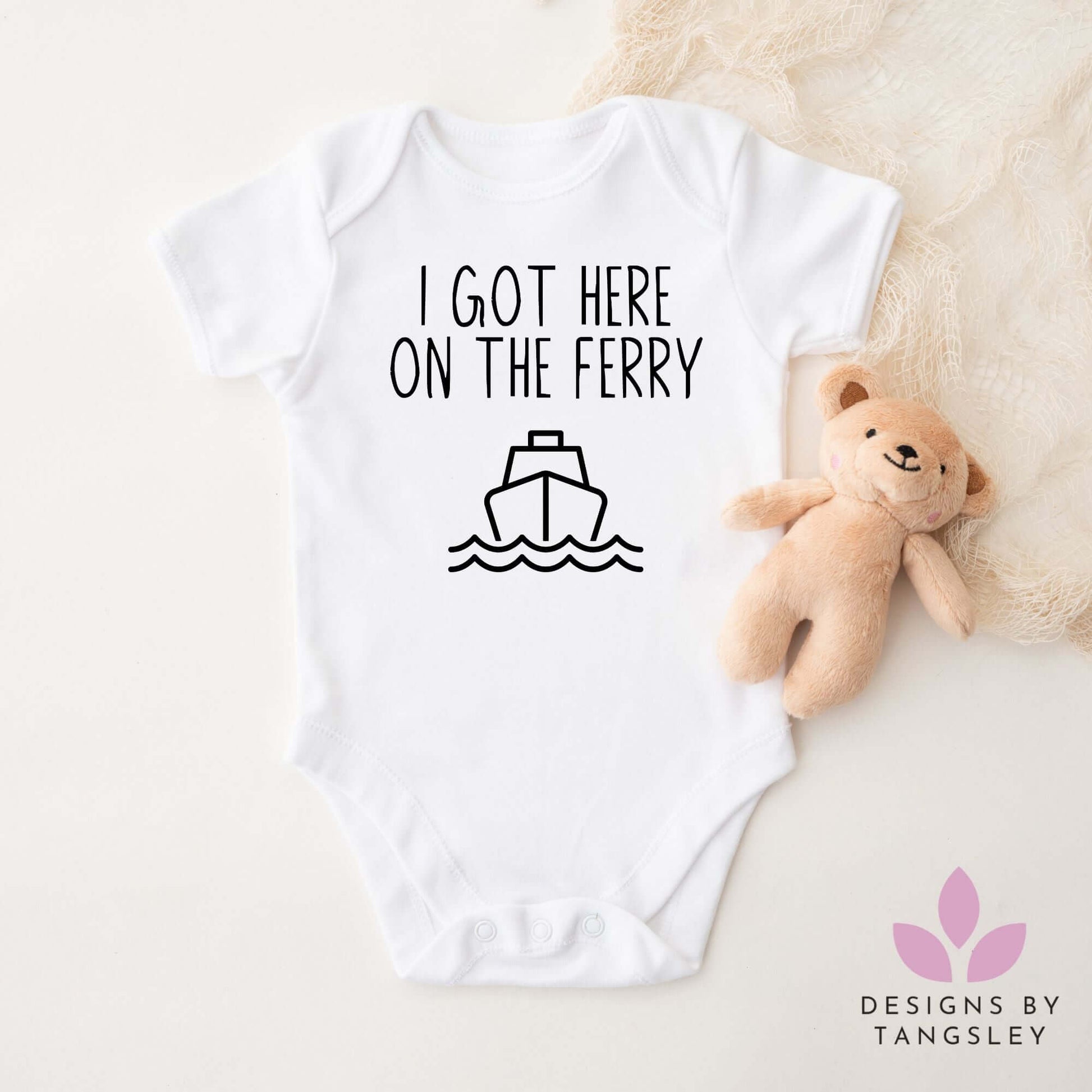 White baby bodysuit with black text "I Got Here On The Ferry" and a simple ferry graphic. Displayed flat with a plush teddy. Handmade by Designs by Tangsley.