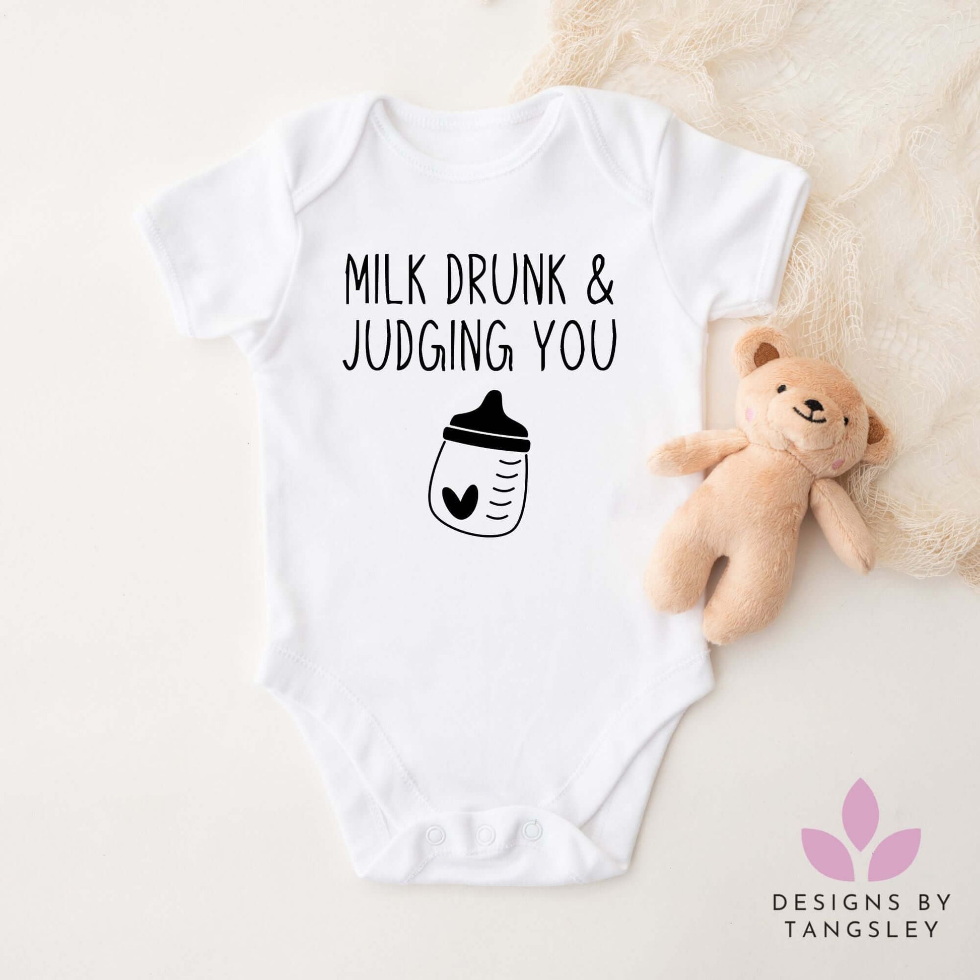 White baby bodysuit with “Milk Drunk & Judging You” text and baby bottle graphic, next to a teddy bear – handmade by Designs by Tangsley.