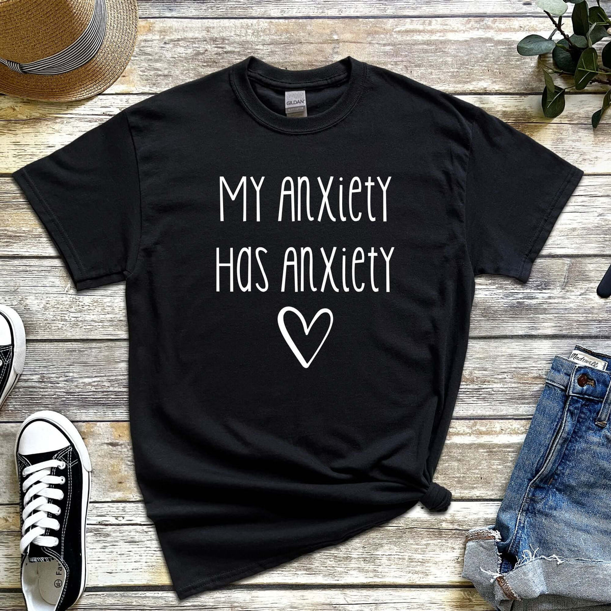 Black t-shirt with “My Anxiety Has Anxiety” in white font, flat lay style with casual accessories, mental health awareness apparel.