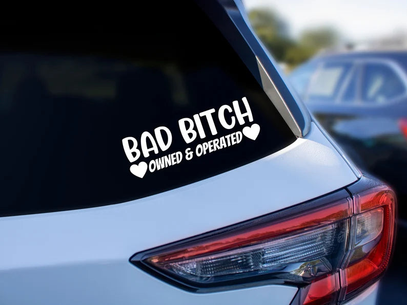 The back of a car. On the window it says "BAD BITCH OWNED & OPERATED" "owned and operated" has a heart on each side.