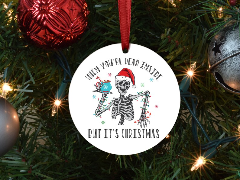 round christmas ornament hanging on a tree. there's a picture of a skeleton in a santa hat drinking hot chocolate. it says "when you're dead inside" above. and "but it's christmas" below it.