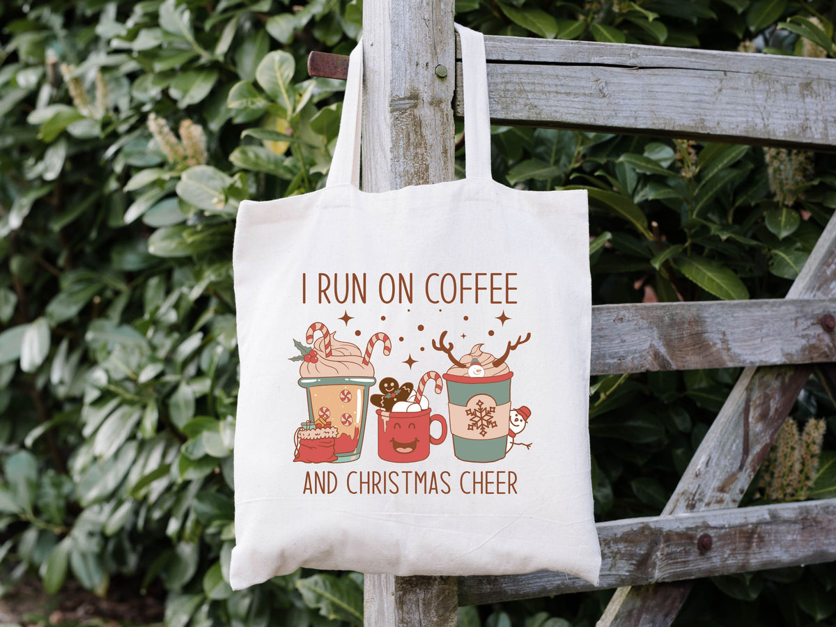 white tote bag hanging on a fence. it says "i run on coffee and christmas cheer" with images of different christmas drinks with cute christmas things like reindeer antlers, gingerbread men, candy canes and snowmen.
