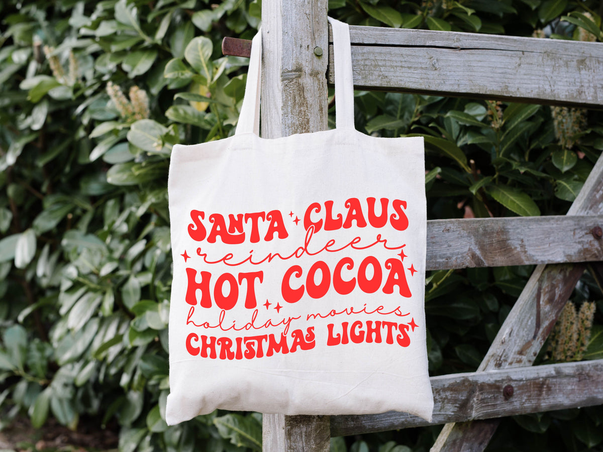 white tote bag hanging on a fence. it says "santa claus, reindeer, hot cocoa, holiday movies, christmas light" in fun fonts in red.