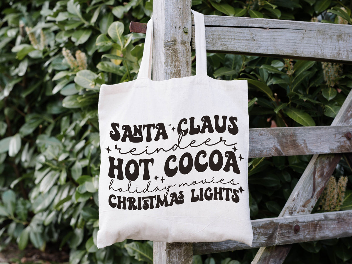 white tote bag hanging on a fence. it says "santa claus, reindeer, hot cocoa, holiday movies, christmas light" in fun fonts in black.