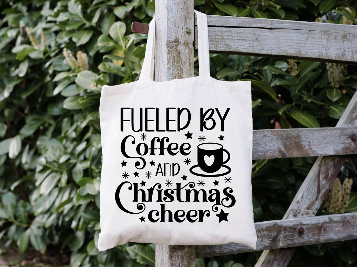 white tote bag hanging on a fence. it says "fueled by coffee and christmas cheer" in fun fonts in black with a coffee cup with a heart on it.