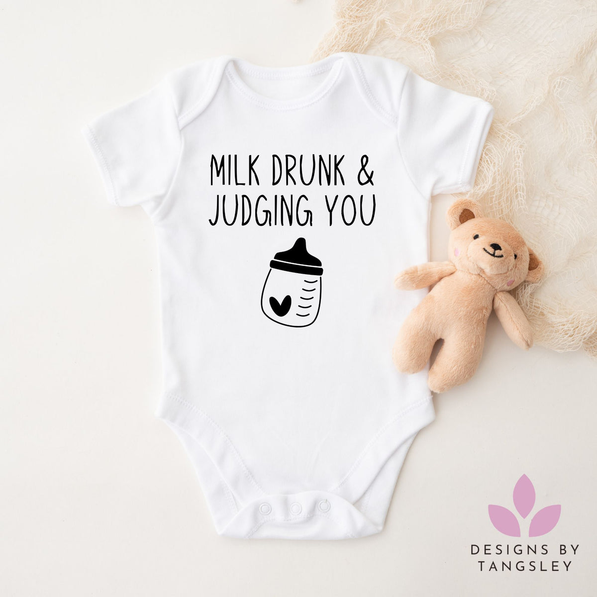 White baby bodysuit with “Milk Drunk & Judging You” text and baby bottle graphic, next to a teddy bear – handmade by Designs by Tangsley.