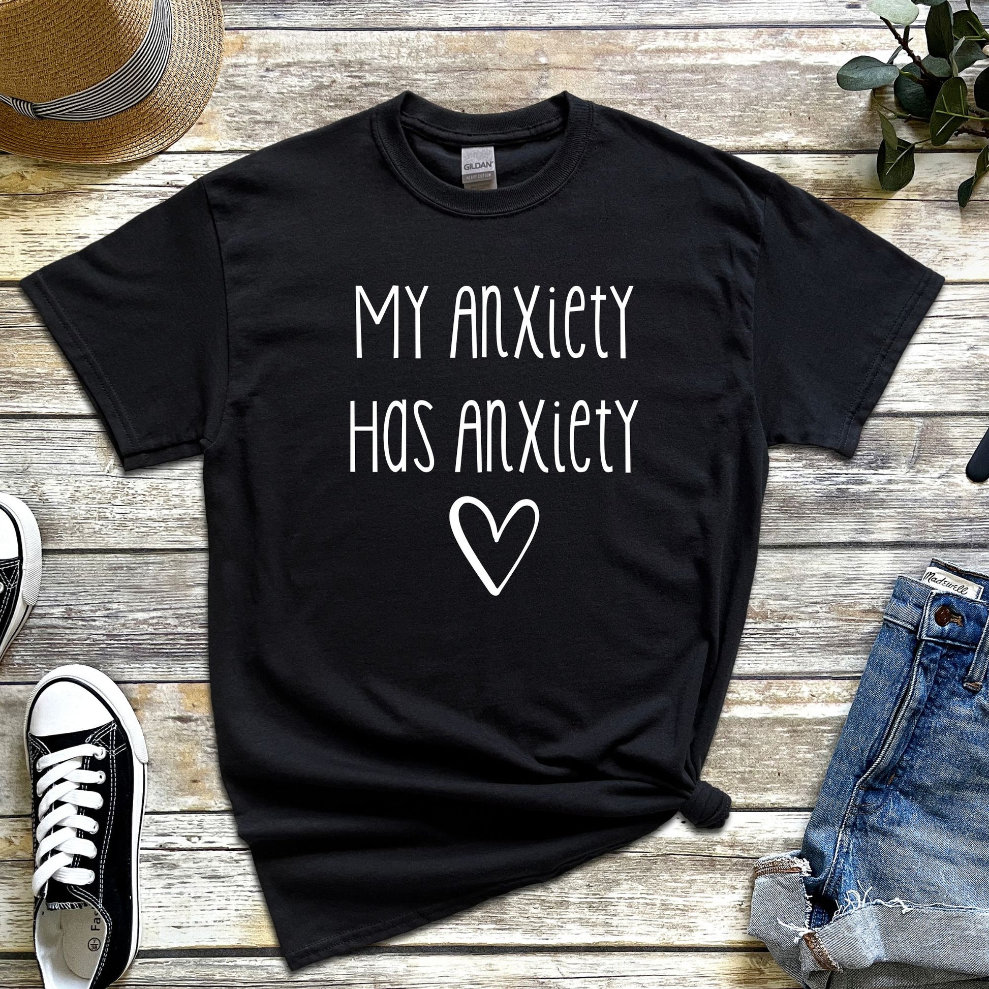 Black t-shirt with “My Anxiety Has Anxiety” in white font, flat lay style with casual accessories, mental health awareness apparel.