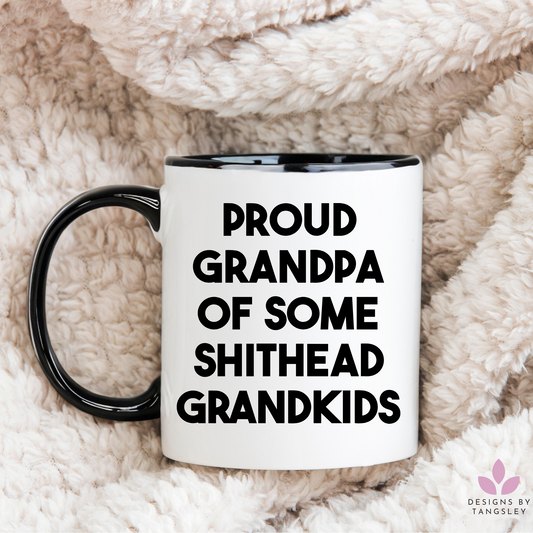 white mug with black handle and black inside. it says proud grandpa of some shithead grandkids in a bold black font.