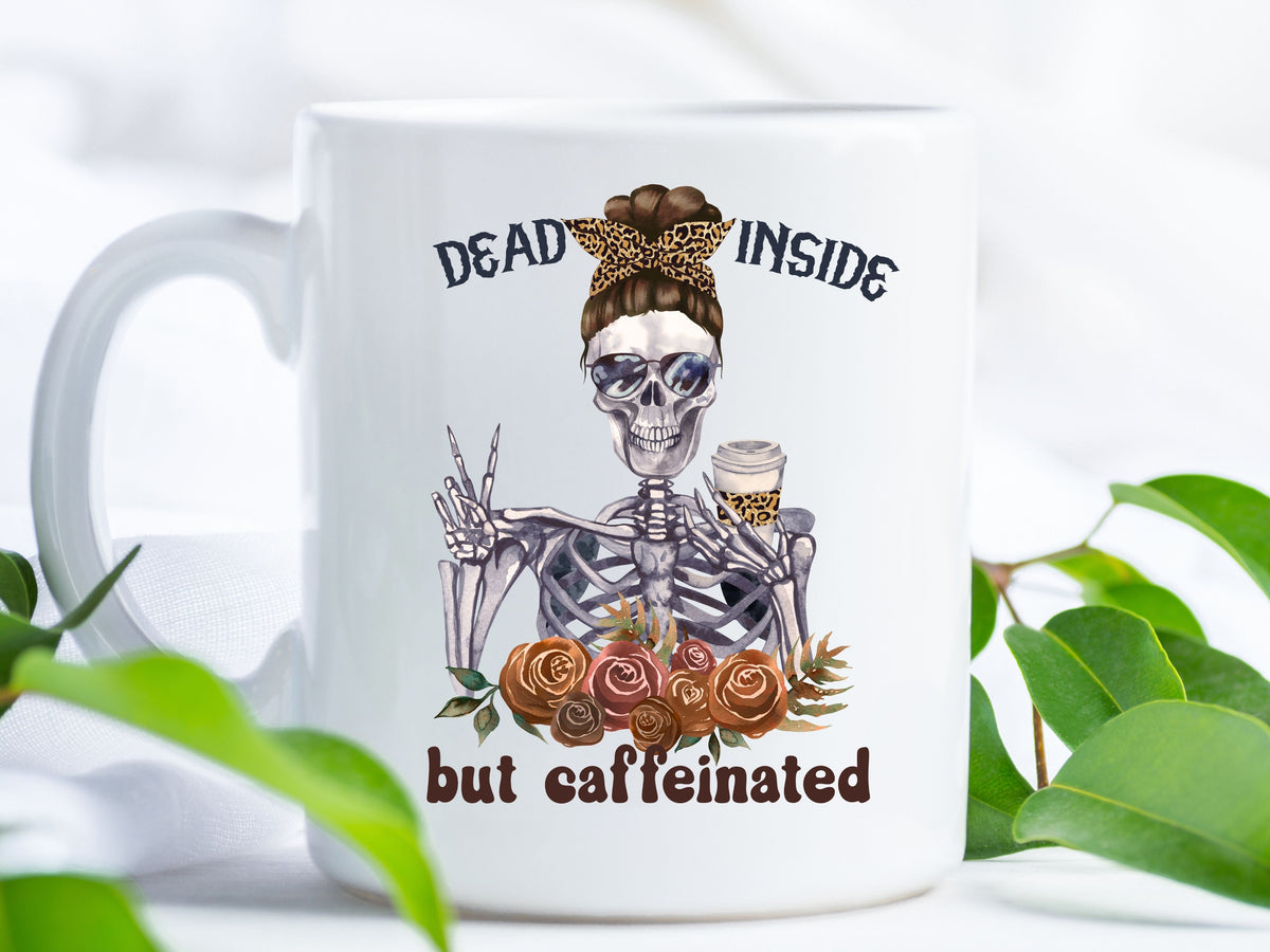 white mug with handle. it has "dead inside but caffeinated" on it with a picture of a skeleton with a messy bun and cheetah print accents giving a peace sign and drinking coffee in sunglasses.